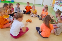 Dzieci siedzą w kole na podłodze, pośrodku leży marchewka na talerzu, w rytm muzyki podają sobie marchewkę z ręki do ręki.