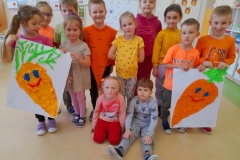 Grupadzieci stoi, dwie dziewczynki I dwóch chłopców trzyma w rękach skończone prace, wyklejone marchewki z bibuły.