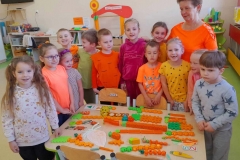 Wokół stołu stoją dzieci z wychowawczynią, na stole leżą marchewki ułożone z różnych Klocków przedszkolnych.