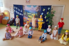 Kilkoro dzieci stoi, recytuje wiersz. Grupa dzieci siedzi na podłodze, na głowach mają opaski: majowe kwiatki. Za nimi baner z napisem: Dzień Rodziny oraz majową łąką.