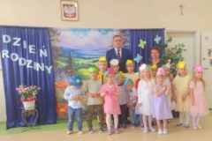 Dzieci wraz z wychowawczynią i wójtem gminy stoją na tle kolorowej tablicy przedstawiającej majową łąkę.