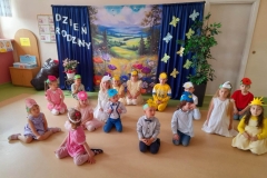 Grupa dzieci siedzi na podłodze, na głowach mają opaski: majowe kwiatki. Za nimi baner z napisem: Dzień Rodziny oraz majową łąką.