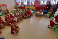Dzieci siedzą w półkolu na podłodze, przed nimi prezenty, słuchają bajki opowiadanej przez Mikołaja