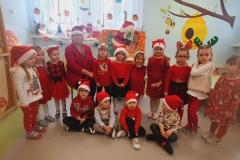 Czworo dzieci siedzi na podłodze. Dziesięcioro stoi tworząc półkole. Pośrodku siedzi pani w czerwonym stroju W tle widać okna.