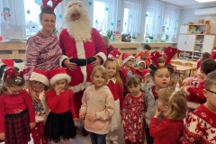 Dzieci stoją w rozsypce na sali, pośród nich Mikołaj a przy nim pani. W tle widać okna.