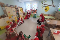 Dzieci siedzą na podłodze tworząc półkole. Posrodku siedzi Mikołaj. W tle widać szafki, stoliki i okna.