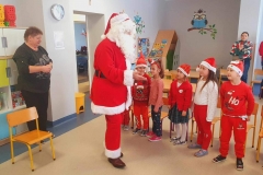 Pięcioro dzieci stoi, patrzy w kierunku Mikołaja. Obok stoi pani ubrana na ciemno.  W tle widać szafki.