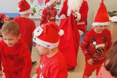 Mikołaj siedzi na krześle. Obok stoją dzieci ubrane w czerwone stroje.