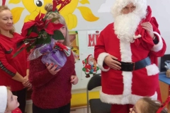 Mikołaj w czerwonym stroju stoi posrodku sali, obok pani która trzyma w rękach kwiatka.