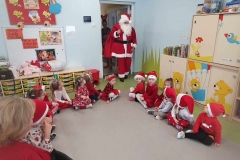 Dzieci ubrane w czerwone stroje siedzą na podłodze tworząc półkole. Mikołaj ubrany w biało- czerwony strój stoi pośrodku dzieci. Z prawej strony stoją  szafki, z lewej widać  tablicę.