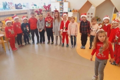 Dzieci w kolorowych strojach stoją obok siebie tworząc półkole. W tle widać szafki i okna.