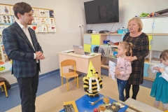 Na zdjęciu są  widoczne 2 dorosłe  osoby oraz dziecko   Dziewczynka trzyma w rękach kartkę. W tle telewizor.