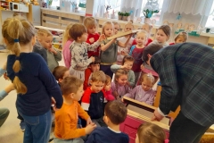 Dzieci zgromadzone wokół stołu, na stole leżą słoiczki z miodem, postacie pszczoły i trutnia. Pan opowiada dzieciom o życiu pszczół.