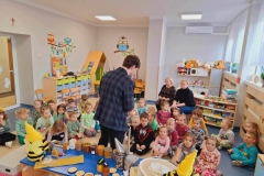 Na stoliku stoją słoiki z żółtym  miodem. Widać  też  pana ubranego w ciemny strój. Przed nim siedzą  na podłodze dzieci, a na krzesełkach 2 panie.