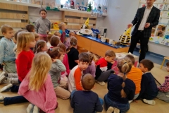 Dzieci zgromadzone wokół stołu na którym leżą miody w słoiczkach. Pan pokazuje dzieciom plaster miodu.