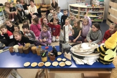 Na stoliku stoją niewielkie sloiki z miodem. Dalej  widać dzieci siedzące w rozsypce na podłodze oraz 4 panie. W tle  są półeczki i szafki z kolorowymi zabawkami.