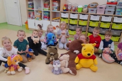 Dzieci ubrane w kolorowe  stroje wraz z panią ubraną  na brązowo siedzą  na podłodze.  Przedszkolaki trzymają  w rączkach kolorowe pluszaki. W tle,  sylweta dużego misia i napis.
