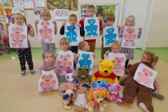 Kilkoro dzieci stoi, przed nimi kilkoro dzieci siedzi na podłodze, w rękach trzymają prace przedstawiające kolorowe Misie. Przed dziećmi ustawione różne pluszaki.