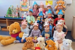 Kilkoro dzieci siedzi na podłodze,  pozostałe stoją za nimi. Przed nimi  leżą  kolorowe pluszowe misie.