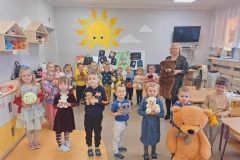 Dzieci stoją w kole, trzymają misie w rączkach.  W tle żółte  słoneczko namalowane na ścianie.