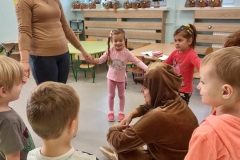  Dzieci wraz z panią stoją w kole. W środku na podłodze siedzi  pani ubrana w brązowy strój.