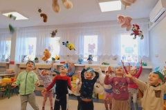 Grupa dzieci ubranych w kolorowe stroje stoi w rozsypce w sali. Mają  uniesione ręce w górę,  gotowe chwycić  wyrzucone wczesniej misie. W tle okna.