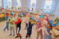 Grupa dzieci ubranych w kolorowe stroje stoi w rozsypce w sali. Mają  uniesione ręce w górę,  gotowe chwycić  wyrzucone wczesniej misie. W tle okna.