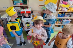 Przed szafkami stoi: 2 chłopców- robotów, dziewczynka w kapeluszu w kwiecistej sukience, z torebką na ramieniu, chłopiec- bateria. Za nimi chłopiec w kapeluszu z folii aluminiowej i dziewczynka w okularach.