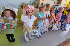 Grupa dzieci w oryginalnych strojach wykonanych z kartonów, serwetek, zakrętek stoi na szaro- zielonej podłodze. Za nimi szafka z książkami.