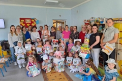 Grupa dzieci ubrana w stroje ekologiczne siedzi lub stoi na podłodze. W rękach trzymają dyplomy I książki. Za nimi stoją mamusie. Na ścianie wisi czerwona tablica z pszczołami.