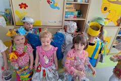 Wanda Sobota 3 dziewczynki w różowych bluzkach stoi obok siebie. Jedna ma na głowie wianek z niebieskich i żółtych kwiatów. Za nimi 2 chłopców w czapkach z folii aluminiowej. Jeden odwrócony plecami z żółtą opaską na głowie.