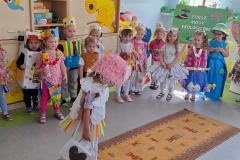 Na wzorzystym chodniku dziewczynka w białej bluzeczce z serwetek, pasiastej spódnicy, różowymi włosami. W ręku trzyma białą torebkę z czarnym sercem. Za nią grupa dzieci w kolorowych strojach.