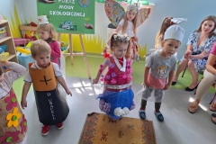 Dzieci ustawione na środku sali: chłopiec- bateria, dziewczynka w czerwono- niebieskiej sukience z kwiatami, chłopiec z białą czapką na głowie. Za nimi dziewczynka- motyl oraz dziewczynka w różowym sweterku. W tle zielona tablica z napisem: Pokaz Mody Ekologicznej.