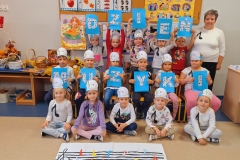 Sala przedszkolna . Dzieci ubrane na kolorowo z opaskami na głowach ustawione przed tablicą . Każde dziecko ma przypiętą do koszulki jedną nutkę . Po lewej stronie widać stoliki na których stoją wiklinowe koszyki z darami przyrody. W tle za dziećmi 2 tablice korkowe z kolorowymi ilustracjami . Przed dziecmi na podłodze na białej prostokątnej płaszczyźnie pięciolinia z kluczem wiolinowym i kolorowymi nutkami, oraz napis DZIEŃ MUZYKI na prostokątnych kartkach papieru w kolorze niebieskim.