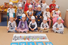 Sala przedszkolna . Dzieci ubrane na kolorowo z opaskami na głowach ustawione przed tablicą . Niektóre z dzieci trzymają w rękach niebieskie prostokątne kartki z białymi literami na których widnieją obrazy graficzne nutek .Litery tworzą napis DZIEŃ MUZYKI.Każde dziecko ma przypiętą do koszulki jedną nutkę. Po prawej stronie stoi osoba dorosła . Po lewej stronie widać stoliki na których stoją wiklinowe koszyki z darami przyrody. W tle za dziećmi 2 tablice korkowe z kolorowymi ilustracjami . Przed dziecmi na podłodze na białej prostokątnej płaszczyźnie pięciolinia z kluczem wiolinowym i kolorowymi nutkami.