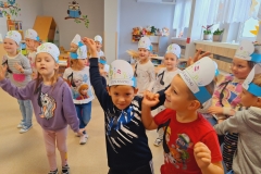 Sala przedszkolna . Dzieci ubrane na kolorowo w luźnej rozsypce z opaskami na głowach tańczą do muzyki przyjmując różne ustawienia. Ad5.Pięć dziewczynek siedzi przy prostokątnym stoliku z opaskami na głowiach. Dziewczynki ukladają czarne nutki na dużej pięciolinii.