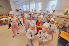 Sala przedszkolna . Dzieci ubrane na kolorowo w luźnej rozsypce z opaskami na głowach tańczą do muzyki przyjmując różne ustawienia. Ad5.Pięć dziewczynek siedzi przy prostokątnym stoliku z opaskami na głowiach. Dziewczynki ukladają czarne nutki na dużej pięciolinii.