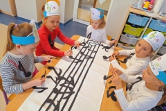 Jedna dziewczynka i czterech chłopców siedzą przy stolikach i układają kompozycje z kolorowych nutek na białych prostokątnych kartkach.