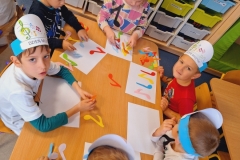 Kilka dziewczynek ubranych na kolorowo i w opaskach na głowach ( 5 ) siedzą przy prostokątnym stoliku.Na środku stolika leżą kolorowe nutki . Przed każdą dziewczynką biała prostokatna kartka. Na jednej z kartek leżą 2 różowe papierowe nutki