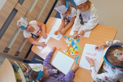 Kilka dziewczynek ubranych na kolorowo i w opaskach na głowach ( 5 ) siedzą przy prostokątnym stoliku.Na środku stolika leżą kolorowe nutki . Przed każdą dziewczynką biała prostokatna kartka. Na jednej z kartek leżą 2 różowe papierowe nutki