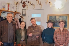 5 osób stoi w półkolu. W środku pan w brązowo- beżowej koszuli w kratę z rękami skrzyżowanymi na brzuchu. Z prawej jego strony pani w ciemnozielonej marynarce. Obok niej pan w szarej koszuli i brązowej kurtce. Z lewej strony pani w granatowym swetrze i zielonym kapeluszu na głowie. Przy niej pani w kraciastej koszuli.  Za nimi na ścianie zawieszone porożami zwierząt.