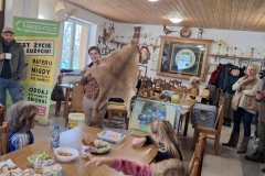 Pani w koszuli w kratkę trzyma w rękach skórę z kangura. Przed przy stole siedzą dzieci. Z boku stoi pan w zielonej kurtce i beżowej czapce z białym kubkiem w ręce. W tyle półka z pucharami w kolorze złotym, bażant, głowa sarny.