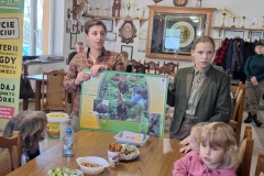 Pani w koszuli w kratkę oraz pani w jasnozielonej koszuli i ciemnozielonej marynarce stoją przy stoliku. W rękach trzymają planszę z obrazkami niedźwiedzia. Przed nimi siedzą dzieci. W tyle stoi pani w kapeluszu, przy stoliku siedzi pan.