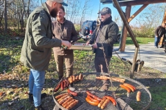Na ruszcie ułożone kiełbaski. Obok grilla stoi 3 panów. Jeden trzyma w rękach srebrną tackę, drugi szczypce z zielonymi uchwytami. W tle drzewa, samochód, fragment drewnianej altanki. Na trawie kolorowe liście.