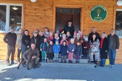 Na schodach przed drewnianym domkiem stoi grupa dzieci. Za dziećmi na tle drzwi myśliwy w odświętnym stroju. Po bokach panie i panowie. Przed grupą klęczy pan w kraciastej koszuli, a przy nim stoi pies myśliwski koloru ciemnobrązowego. Na ścianie domu wisi zielona tablica w kształcie koła z napisem: Koło Łowieckie " Trop" Wietrzychowice.