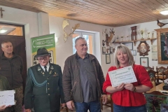 Pani w dżinsach I czerwonej bluzce trzyma w rękach bon podarunkowy. Obok niej stoi pan w szarej koszuli i brązowej kurtce. Przy nim pan w stroju myśliwskim. Za nimi tablica z napisem Argo- Film.