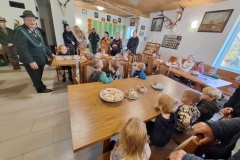 Pan w stroju myśliwego: czarne spodnie, biała koszula, ciemnozielona marynarka z zielonym sznurem i srebrnymi guzikami , zielony kapelusz na głowie- stoi na szarej podłodze. Przed nim przy 3 drewnianych, prostokątnych stolikach siedzą dzieci w kolorowych ubraniach. Z boku stoją 4 panie i 2 panów. Na ścianie wisi obraz i poroża  zwierząt.