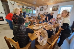 Przy 3 drewnianych, prostokątnych stołach siedzą dzieci. Wśród nich 8 pań i 1 pan. Na stole talerze z rogalikami, ciasteczkami, cukierkami, paluszkami, precelkami. Przed każdym dzieckiem talerzyk z kiełbasą i kubek z herbatą.