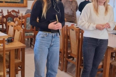 Pani w jasnych dżinsach i czarnej bluzce stoi na szarej podłodze. Przed nią biały pojemnik z żółtymi naklejkami. Obok niej pani w ciemnych dżinsach i białym swetrze.