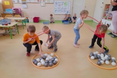 Dwóch chłopców I dwie dziewczynki są w dwóch obręczach, przenoszą piłeczki na drugą stronę sali. Pod ścianą siedzi reszta dzieci, kibicuje.